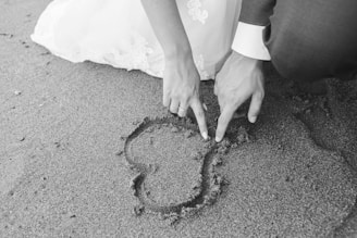 grayscale photography of man and woman draw heart on sand