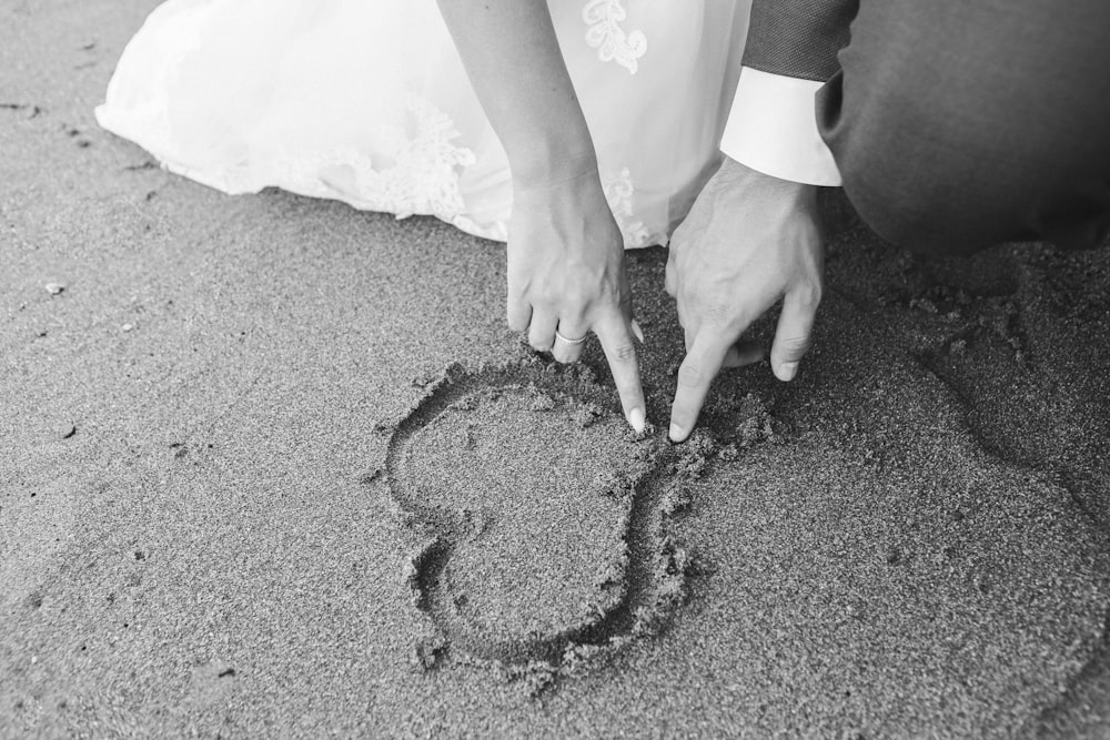 grayscale photography of man and woman draw heart on sand