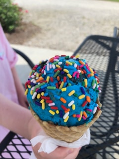 A bright blue ice cream cone covered with colorful sprinkles. The ice cream is held by a hand atop a waffle cone, and a napkin is wrapped around the base. The background includes a metal outdoor table and a garden area with greenery and flowers.