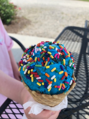 A bright blue ice cream cone covered with colorful sprinkles. The ice cream is held by a hand atop a waffle cone, and a napkin is wrapped around the base. The background includes a metal outdoor table and a garden area with greenery and flowers.