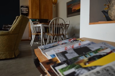 A cozy living space featuring a green upholstered armchair and a wooden dining set in the background. A stack of colorful magazines is prominently placed on a table in the foreground. The room includes wooden cabinets, decorative items, and a wall-mounted artwork.