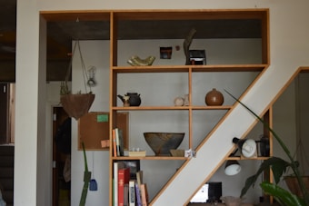 A modern wooden shelf structure filled with various decorative items, including pottery, books, and a hanging plant. The shelf is split into multiple compartments creating an asymmetric design. A large plant is visible at the bottom right, and the lighting is soft, giving the room a cozy feeling.