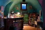 A cozy, dimly-lit wine cellar features shelves lined with bottles of wine and spirits. A small wooden table displays various glassware under a warm, glowing table lamp, creating a rustic and inviting atmosphere. The turquoise walls add a unique charm to the room, with a small window casting subdued natural light. Decorative elements include framed pictures and vintage wine crates.