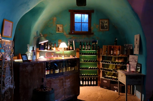 A cozy, dimly-lit wine cellar features shelves lined with bottles of wine and spirits. A small wooden table displays various glassware under a warm, glowing table lamp, creating a rustic and inviting atmosphere. The turquoise walls add a unique charm to the room, with a small window casting subdued natural light. Decorative elements include framed pictures and vintage wine crates.