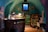 A cozy, dimly-lit wine cellar features shelves lined with bottles of wine and spirits. A small wooden table displays various glassware under a warm, glowing table lamp, creating a rustic and inviting atmosphere. The turquoise walls add a unique charm to the room, with a small window casting subdued natural light. Decorative elements include framed pictures and vintage wine crates.
