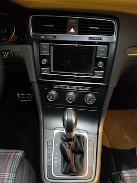 The interior of a car dashboard features a central touchscreen display surrounded by control buttons. Below the screen are air conditioning controls and a gear shift with black leather and red stitching. To the side, plaid fabric is visible on the seat.