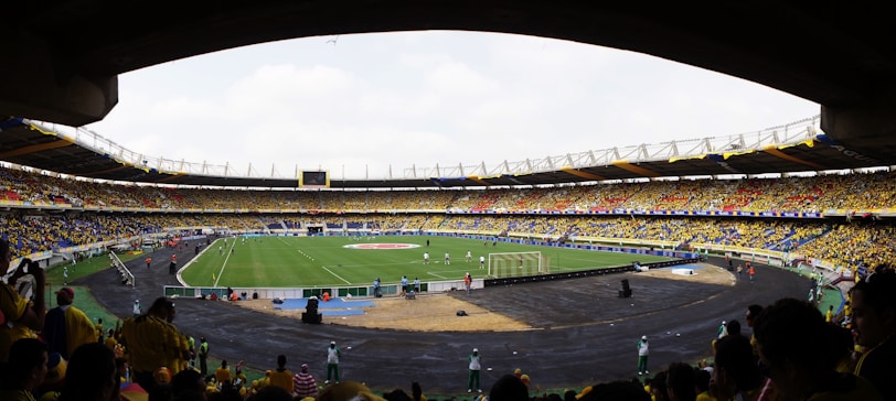 A large football stadium filled with spectators, with a green field in the center where a match is taking place. The crowd is mostly wearing yellow shirts, with some people wearing other colors. The stadium has large stands with seats, and numerous advertising banners are visible. The atmosphere appears lively and energetic.