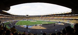 A large football stadium filled with spectators, with a green field in the center where a match is taking place. The crowd is mostly wearing yellow shirts, with some people wearing other colors. The stadium has large stands with seats, and numerous advertising banners are visible. The atmosphere appears lively and energetic.