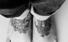 A black and white photograph of two tattooed feet resting side by side, with rolled-up denim jeans. The visible tattoos include a rose with the word 'Passion' written across it and another tattoo with a floral design and text.