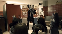 A magician performing a trick in front of a group of children, with some of them seated and others standing. The setting appears to be indoors with red, checked walls and decorative plates. A man in a white shirt and red apron is standing beside the magician, observing the performance.