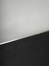 A simple interior corner with a white wall and a dark floor separated by a thin metallic strip. The wall has light scratches visible upon close inspection.