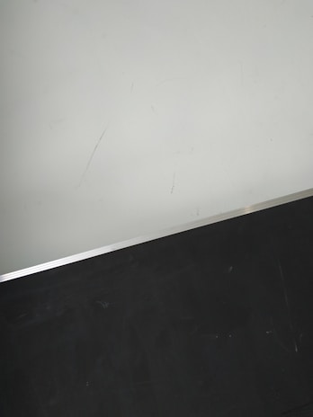 A simple interior corner with a white wall and a dark floor separated by a thin metallic strip. The wall has light scratches visible upon close inspection.