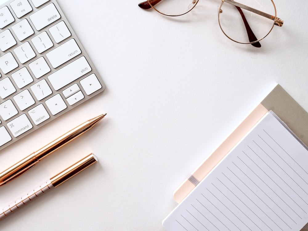 Pen near black lined paper, laptop keyboard, and eyeglasses. Photo by Jess Bailey via Unsplash. 