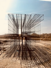 A construction site with large, rectangular steel rebar structures placed on a dirt path. The backdrop includes a clear sky and barren, earthy mounds. Shadows of the rebar create intricate patterns on the ground.