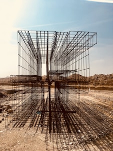 A construction site with large, rectangular steel rebar structures placed on a dirt path. The backdrop includes a clear sky and barren, earthy mounds. Shadows of the rebar create intricate patterns on the ground.