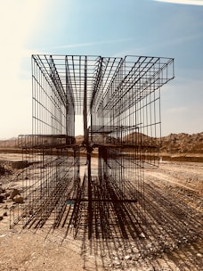 A construction site with large, rectangular steel rebar structures placed on a dirt path. The backdrop includes a clear sky and barren, earthy mounds. Shadows of the rebar create intricate patterns on the ground.