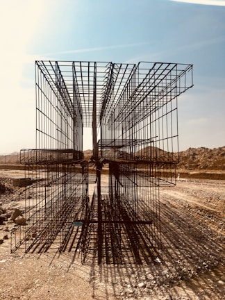 A construction site with large, rectangular steel rebar structures placed on a dirt path. The backdrop includes a clear sky and barren, earthy mounds. Shadows of the rebar create intricate patterns on the ground.