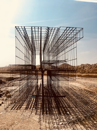 A construction site with large, rectangular steel rebar structures placed on a dirt path. The backdrop includes a clear sky and barren, earthy mounds. Shadows of the rebar create intricate patterns on the ground.