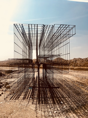 A construction site with large, rectangular steel rebar structures placed on a dirt path. The backdrop includes a clear sky and barren, earthy mounds. Shadows of the rebar create intricate patterns on the ground.