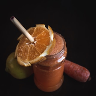 A glass jar filled with an orange-colored smoothie, garnished with a half slice of orange. A straw is inserted into the smoothie. Beside the jar, a carrot and a green apple are placed on a dark background.