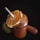 A glass jar filled with an orange-colored smoothie, garnished with a half slice of orange. A straw is inserted into the smoothie. Beside the jar, a carrot and a green apple are placed on a dark background.