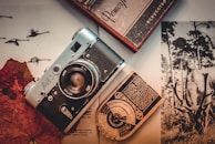 A vintage camera lies on top of an open magazine featuring various black-and-white images, including one of a deer with antlers. Next to the camera is a retro light meter with Cyrillic script and a hardcover book. The setting is enhanced by a sepia tone, giving it a nostalgic feel.