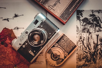 A vintage camera lies on top of an open magazine featuring various black-and-white images, including one of a deer with antlers. Next to the camera is a retro light meter with Cyrillic script and a hardcover book. The setting is enhanced by a sepia tone, giving it a nostalgic feel.