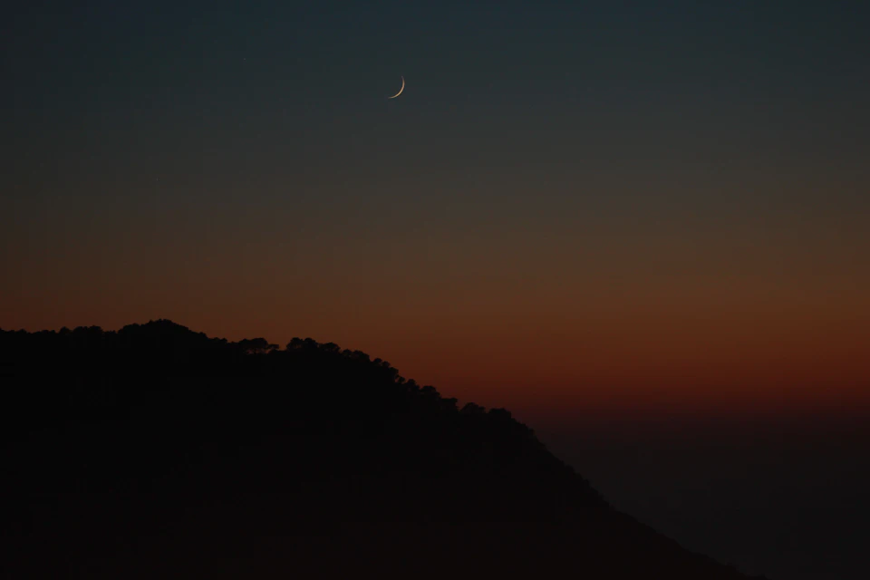 The image depicts a serene night sky with a crescent moon partially obscured by clouds, casting a soft glow over the landscape below. The moon is positioned on the right side of the frame, its light illuminating the scene and creating a sense of tranquility. Below, a hill blanketed in trees stretches across the horizon, adding depth to the image. The sky transitions from deep blue at the top to a lighter shade towards the bottom, with the crescent moon appearing as a small white dot against this backdrop.

The overall composition of the image is balanced and harmonious, with the moon's position on the right side drawing the viewer's attention while the trees in the background provide a sense of depth and scale. The colors used are predominantly dark and light blue, creating a soothing atmosphere that complements the peacefulness of the scene.