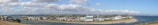 A panoramic view of a coastal town with industrial buildings and warehouses adjacent to a river. The horizon is marked by rolling hills, and a clear blue sky is adorned with scattered fluffy clouds. The waterway separates the town from a sandy strip of land leading into the sea.