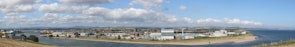 A panoramic view of a coastal town with industrial buildings and warehouses adjacent to a river. The horizon is marked by rolling hills, and a clear blue sky is adorned with scattered fluffy clouds. The waterway separates the town from a sandy strip of land leading into the sea.