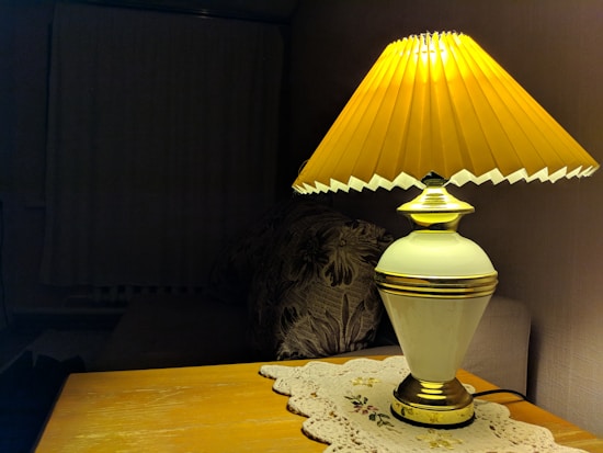A table lamp with a golden-yellow pleated shade emits a warm glow, sitting on a wooden surface partially covered by an embroidered doily. The background is dimly lit, revealing a floral pattern cushion on a sofa.