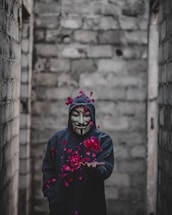 A person wearing a dark hoodie and a white mask with a stylized mustache and goatee stands in an alley with rough stone walls. Red rose petals appear to be floating or scattered around the person, adding a stark contrast to the muted background.