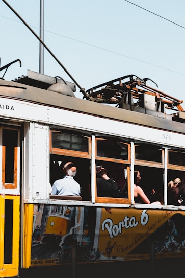 A vintage tram with a yellow and white exterior is moving along a track. Several passengers are visible inside, sitting and looking out the window. The advertisement on the tram features colorful artwork and the text 'Porta 6'. The tram's machinery and overhead wires are visible above.