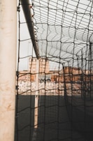 A close-up view of a soccer goalpost and net, with a cityscape visible in the background. The net is prominently in focus, displaying the intricate pattern of the mesh. The setting appears urban, with buildings and a fence visible behind the goal.