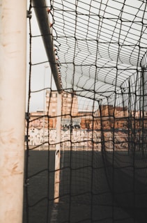 A close-up view of a soccer goalpost and net, with a cityscape visible in the background. The net is prominently in focus, displaying the intricate pattern of the mesh. The setting appears urban, with buildings and a fence visible behind the goal.