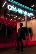 A person stands inside a stylish store, with a neon sign reading 'Off-White' providing a modern and vibrant feel. Clothing items are displayed on racks along the wall, and the store's interior is illuminated with bright, contemporary lighting.