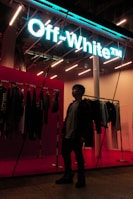 A person stands inside a stylish store, with a neon sign reading 'Off-White' providing a modern and vibrant feel. Clothing items are displayed on racks along the wall, and the store's interior is illuminated with bright, contemporary lighting.