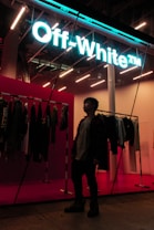 A person stands inside a stylish store, with a neon sign reading 'Off-White' providing a modern and vibrant feel. Clothing items are displayed on racks along the wall, and the store's interior is illuminated with bright, contemporary lighting.