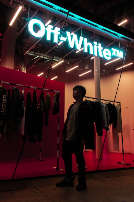 A person stands inside a stylish store, with a neon sign reading 'Off-White' providing a modern and vibrant feel. Clothing items are displayed on racks along the wall, and the store's interior is illuminated with bright, contemporary lighting.