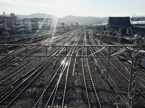 A complex network of railway tracks stretches into the distance under a clear sky, intersected by numerous overhead power cables. Buildings and hills are visible in the background, while sunlight creates a reflection on the rails, adding a dynamic visual element.