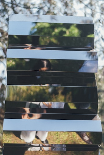 An abstract reflection of a person created by a series of horizontal mirrored panels. The surroundings include greenery, possibly trees or grass, with sunlight casting natural highlights. The reflection appears distorted due to the uneven arrangement of the mirrors.