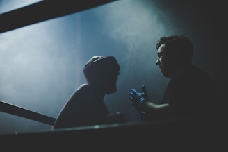Two individuals are in a dimly lit setting, one wearing boxing headgear, suggesting a boxing or athletic context. One person appears to be instructing or motivating the other. The atmosphere is intense with smoke or mist adding dramatic tension.