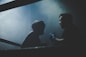 Two individuals are in a dimly lit setting, one wearing boxing headgear, suggesting a boxing or athletic context. One person appears to be instructing or motivating the other. The atmosphere is intense with smoke or mist adding dramatic tension.