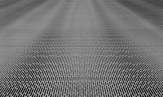 A close-up of a metal mesh structure with intricate interlocking patterns forming a grid-like appearance. The surface is composed of repetitive linear elements creating a sense of depth and complexity.