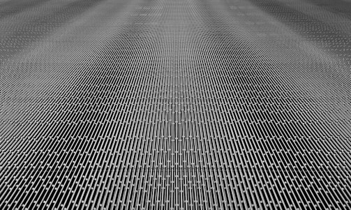 A close-up of a metal mesh structure with intricate interlocking patterns forming a grid-like appearance. The surface is composed of repetitive linear elements creating a sense of depth and complexity.