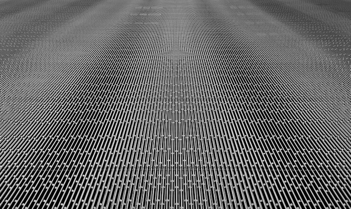 A close-up of a metal mesh structure with intricate interlocking patterns forming a grid-like appearance. The surface is composed of repetitive linear elements creating a sense of depth and complexity.