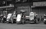 A street scene featuring several makeshift tricycles parked in front of closed shops. People are sitting and resting on the tricycles, with one individual holding an umbrella. The area appears to be a marketplace or urban setting.