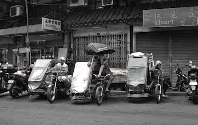 A street scene featuring several makeshift tricycles parked in front of closed shops. People are sitting and resting on the tricycles, with one individual holding an umbrella. The area appears to be a marketplace or urban setting.