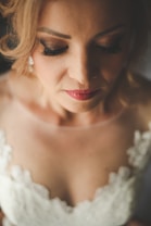 A woman with closed eyes, wearing makeup with prominent eyelashes and red lipstick. She has blonde hair styled in an updo and is dressed in a lace-adorned white garment, likely a wedding dress.