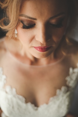 A woman with closed eyes, wearing makeup with prominent eyelashes and red lipstick. She has blonde hair styled in an updo and is dressed in a lace-adorned white garment, likely a wedding dress.